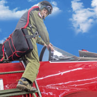 man climbs in small red aircraft wearing army green warbird seat parachute