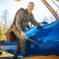 man climbs in vintage aircraft wearing army green warbird seat parachute